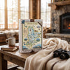 A 1000-piece jigsaw puzzle box featuring a illustrated map of Grand Teton National Park stands on a rustic wooden table in a cozy living room setting. The scene includes a steaming mug on a book, a cream throw blanket, and black binoculars in the foreground, with a stone fireplace and large windows in the background.