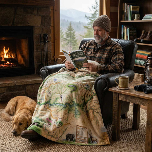Man reading a book in a cozy living room with a fireplace and a dog lying on the floor. He has a Great Smokies blanket on him.