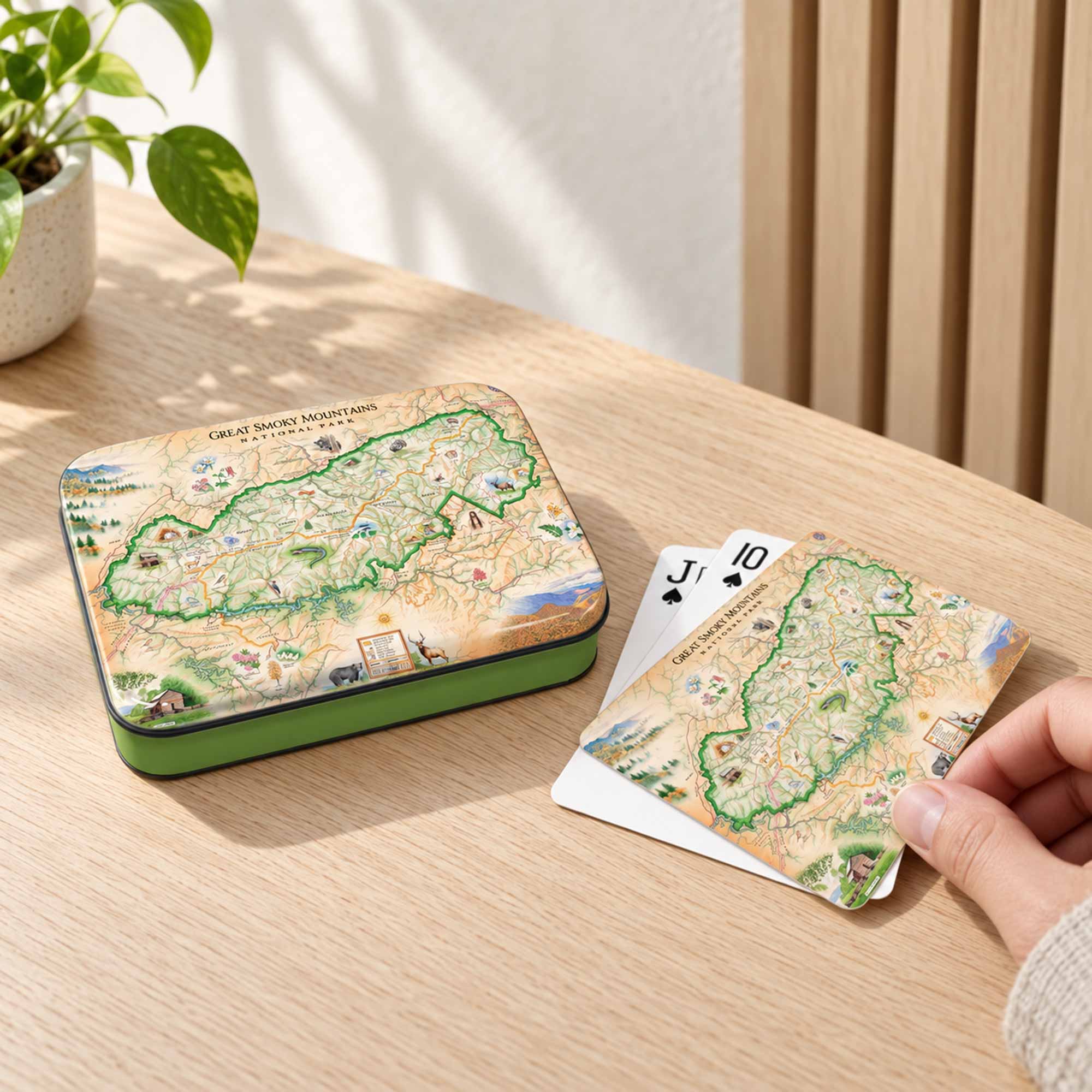 A decorative map-themed playing card tin and matching cards on a light wood table, with a hand holding a card. A potted plant and slatted wall are visible in the background.