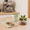 A tall ceramic coffee mug with a green interior and a vintage-style map print on the exterior, styled on a light wood table with a compass, map, coffee beans, and a small plant sprig.