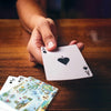 A close-up of a hand holding an Ace of Spades playing card over a wooden table, with other illustrated cards partially visible in the foreground.