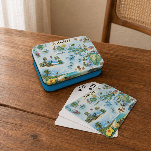 A small illustrated tin of playing cards and a fan of matching cards on a wooden table. The cards and tin feature a tropical Hawaii map design with blue, green, beige, and yellow tones, and the cards have white backs.
