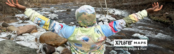 A person holding their hands up in the air. She is standing next to a river in the winter. She is wearing a Missoula map hoodie. 