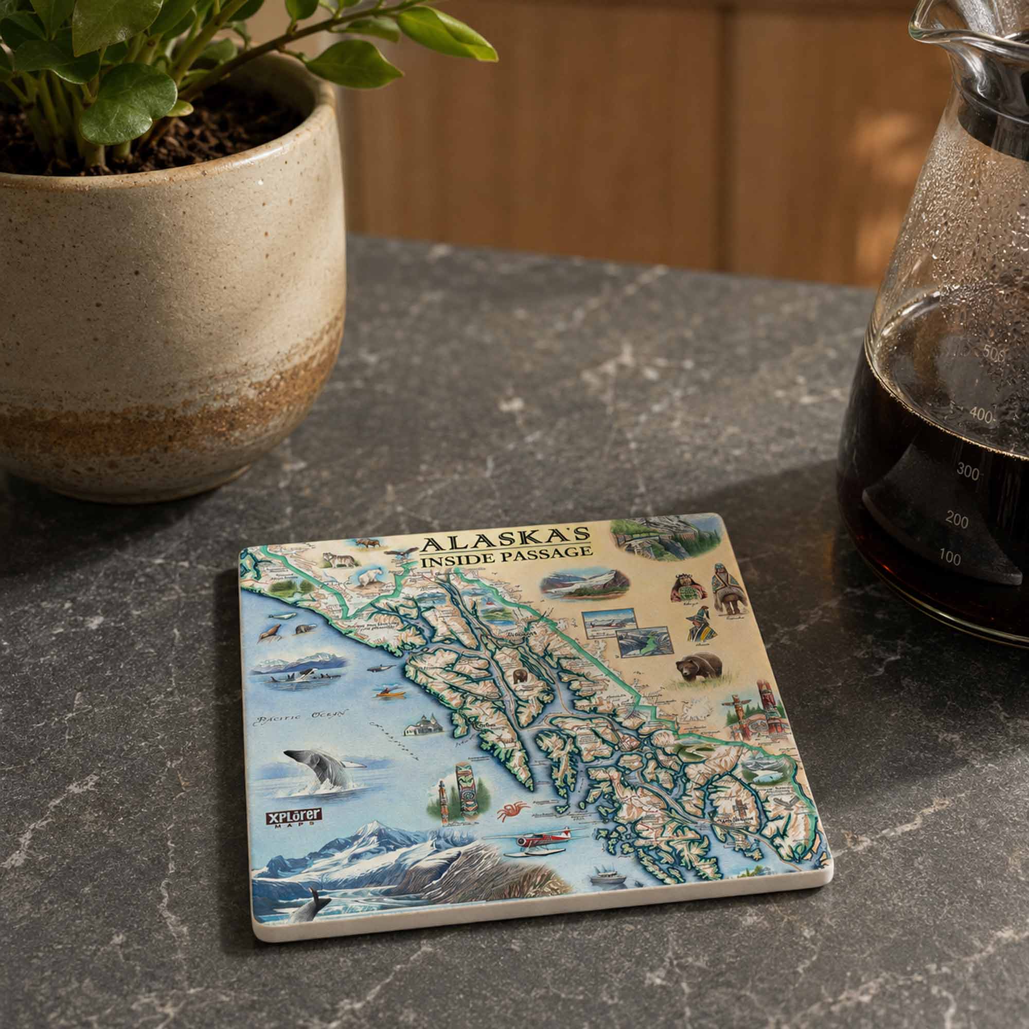 A square ceramic coaster featuring a colorful illustrated map of Alaska's Inside Passage, styled on a dark gray marble countertop next to a potted plant and a glass carafe of coffee.