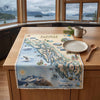 A printed Alaska interior landscape table runner or placemat laid across a wooden kitchen island, with a ceramic mug, plate, and wooden spoon nearby and large windows showing a mountain lake view in the background.