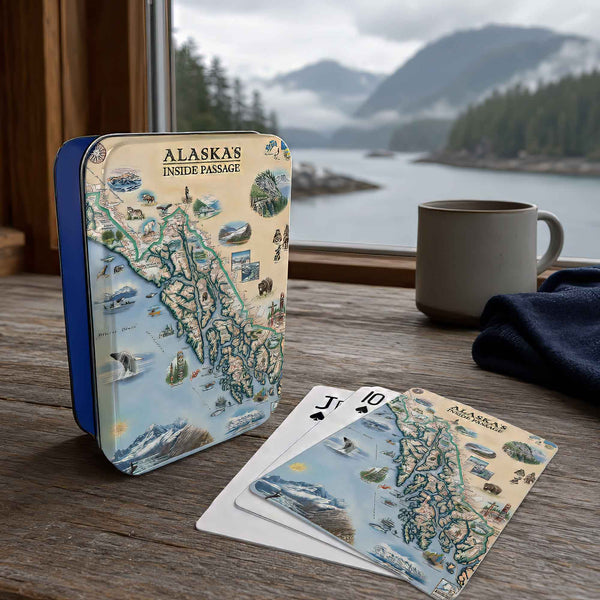 Alaska-themed playing cards and a matching tin box displayed on a wooden table in front of a window with a mountain lake view. A gray mug and dark cloth sit nearby.