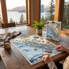 A person is assembling a detailed Alaska map jigsaw puzzle on a rustic wooden table, with the puzzle box standing nearby. The scene is set in a cozy room with large windows overlooking a scenic lake and mountains, with a mug, plant, and blanket on the table.