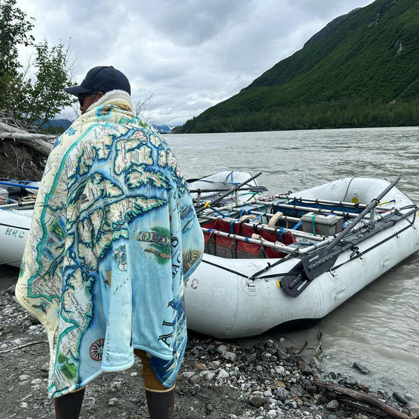 Person wrapped in a Inside Passage blanket standing by a river with rafts and mountains in the background