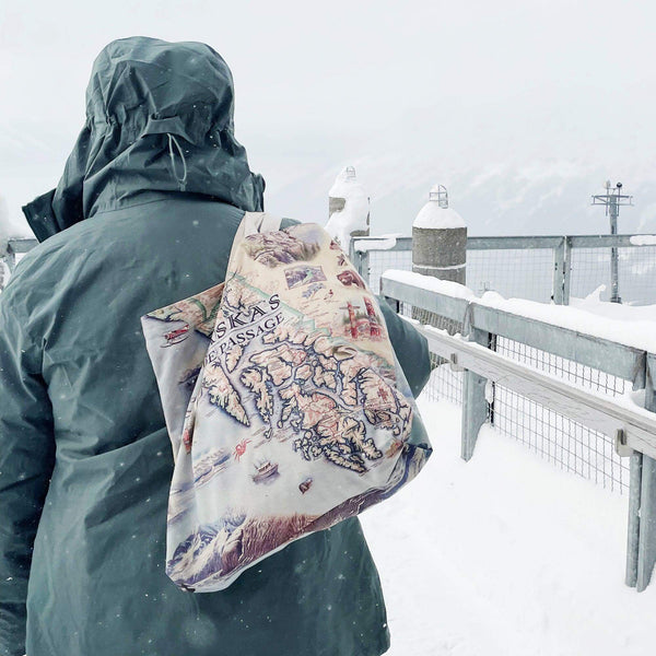 A person wearing a green jacket with a hood up walking over a bridge in the snow. The person is carrying an Alaska Inside Passage canvas tote bag by Xplorer Maps.