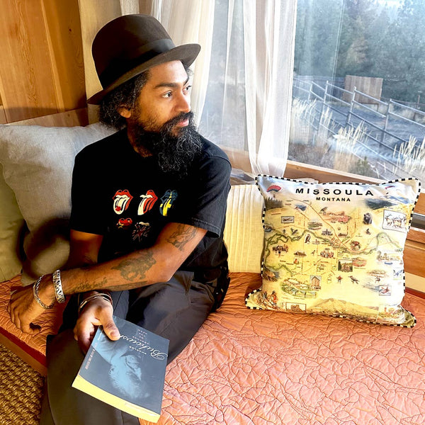 Man sitting in a cozy nook, holding a book and gazing out a window, next to a Missoula, Montana map throw pillow featuring hand-drawn illustrations of local landmarks and wildlife.