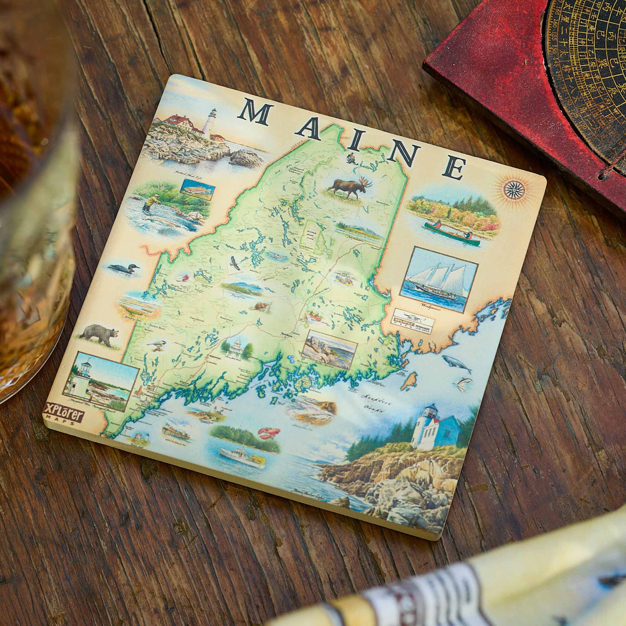 A square coasters featuring a vintage-style illustrated map of Maine, displayed on a rustic wooden tabletop next to a glass and a compass.