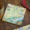 A square coasters featuring a vintage-style illustrated map of Maine, displayed on a rustic wooden tabletop next to a glass and a compass.