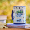 Mug with 'MAINE' design featuring nature scenes on a book with a blurred natural background