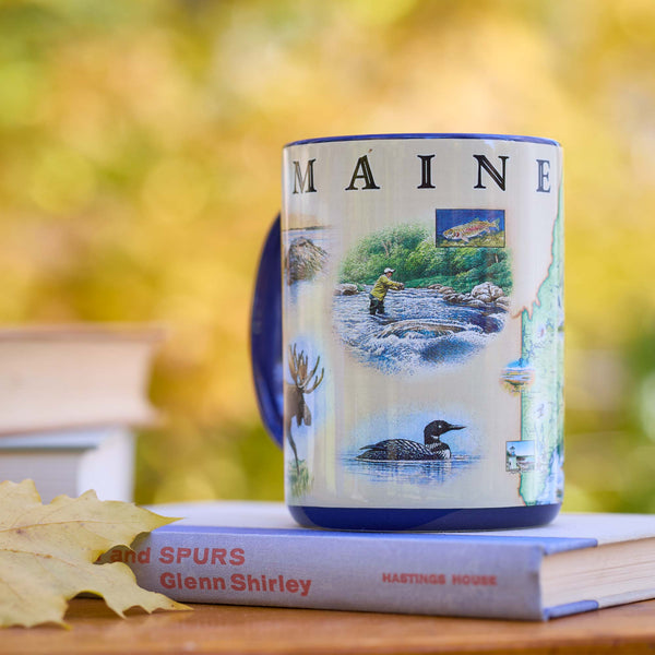 Mug with 'MAINE' design featuring nature scenes on a book with a blurred natural background