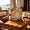 A 10000-piece jigsaw puzzle box featuring an antique-style map of Mallorca, styled on a wooden table with a rolled map, sunglasses, a glass of white wine, and a hat in a sunny outdoor patio setting.