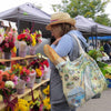 A woman at a farmers market next to fresh-cut flowers. She is carrying a Michigan canvas tote bag by Xplorer Maps with a baguette inside.