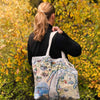 A woman viewed from behind carrying a large canvas tote bag over her shoulder, standing outdoors in front of a background of yellow and green autumn foliage. The tote bag features a detailed map illustration of Michigan with various local landmarks and illustrations.
