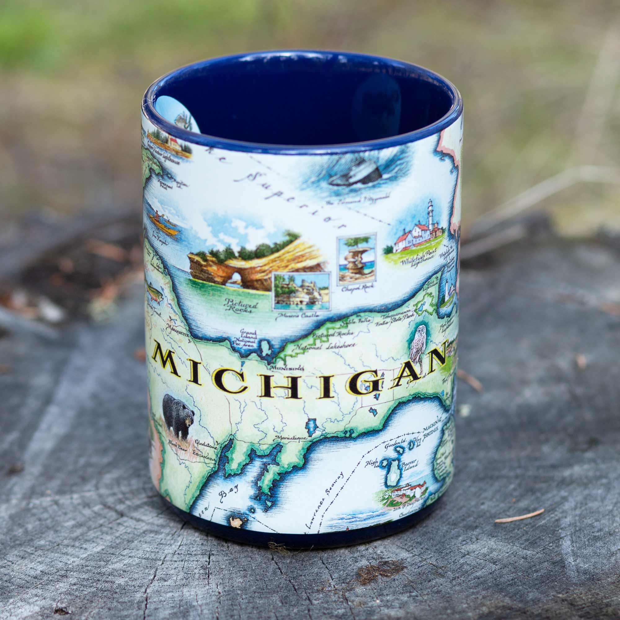 Michigan State Map Ceramic Mug sitting on a log in a forest. The cup features moose, elk, bear, great lakes, lighthouse, fish, Kalamazoo windmill, Detroit, Lansing, Stevie Wonder, Ford Model T, cherries, wolverine, loons, and owls. 