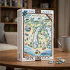 A 1000-piece jigsaw puzzle box featuring a colorful illustrated map of Michigan, displayed on a wooden table with a few loose puzzle pieces in front. A blurred living room background with a bookshelf and a mug is visible.