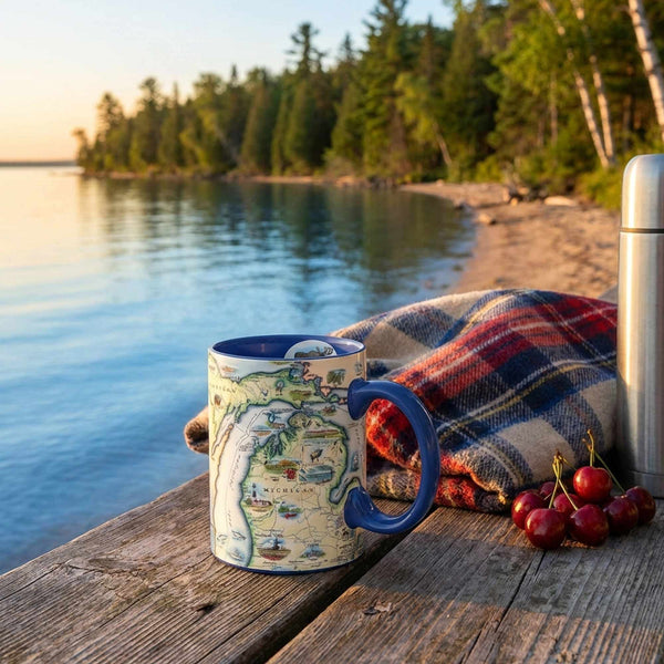Michigan Mug with map design, plaid blanket, and cherries on a wooden dock by a lake.