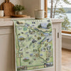 A Minnesota state map print draped over a kitchen counter, featuring illustrated landmarks and local scenes. A ceramic mug sits on the counter above, with a bright kitchen and window in the background.