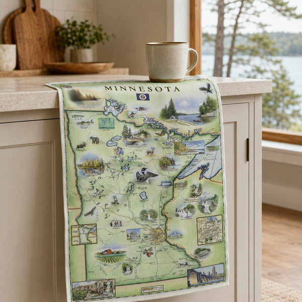 A Minnesota state map print draped over a kitchen counter, featuring illustrated landmarks and local scenes. A ceramic mug sits on the counter above, with a bright kitchen and window in the background.