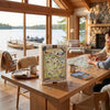 A 1000-piece Minnesota jigsaw puzzle set displayed on a wooden table in a cozy, wood-paneled cabin with a large window overlooking a lake and dock. Two people are seated at the table, working on assembling another puzzle.