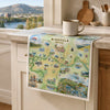 A square kitchen towel featuring a colorful illustrated map of Missoula, Montana, draped over a white kitchen cabinet. The towel is styled in a bright kitchen setting with a ceramic mug on the counter, a scenic window view in the background, and wooden kitchen decor.
