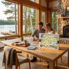A couple sits at a wooden table in a bright cabin with large windows overlooking a lake, working on a Missoula jigsaw puzzle. The puzzle box is prominently displayed on the table, along with mugs, a tray, and a stone fireplace in the background.