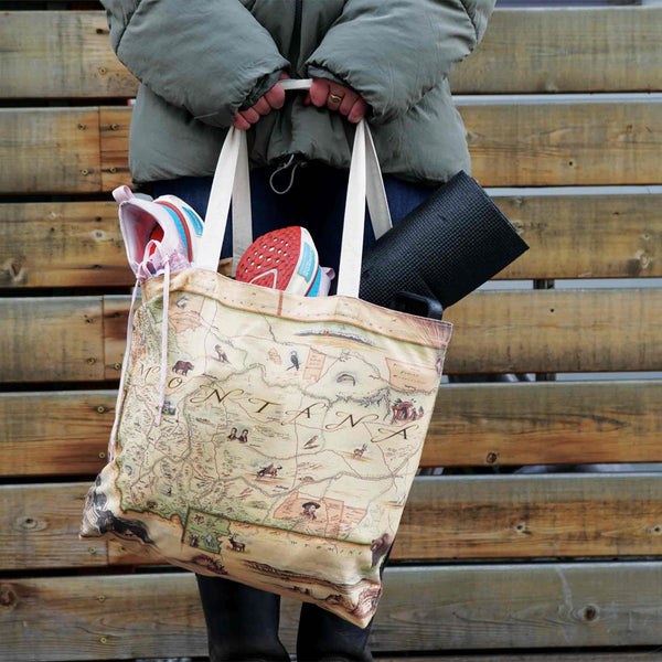 Women by wood fence with Montana Canvas Tote Bag, carrying yoga mat and shoes. Map showcases Sacajawea, Lewis & Clark, Yellowstone, Glacier National Park, Flathead Lake, wildlife like grizzly bear, bald eagle, elk, and cities such as Missoula, Bozeman, Helena, Whitefish.