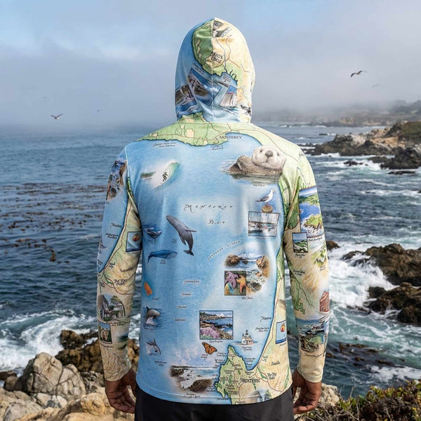 Person wearing a sun hoodie with a map and nature-themed design standing on a rocky coastline.