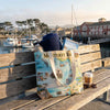 Tote bag with Monterey Bay design on a wooden bench by a waterfront with boats and buildings.