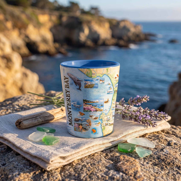 A ceramic mug printed with a colorful Monterey Bay map and coastal imagery, set on a folded linen cloth with sea glass and lavender sprigs on a rocky beach with the ocean and cliffs blurred in the background.