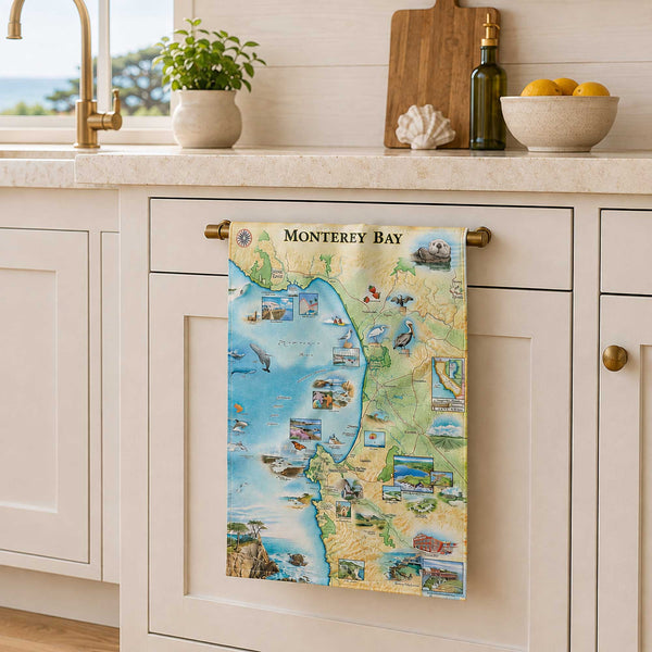A decorative kitchen towel featuring a colorful illustrated map of Monterey Bay, hanging from a brass rack on a white kitchen cabinet. The kitchen setting includes a sink, countertop with a plant, cutting board, and bowl of lemons.
