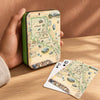 A hand holds a green metal tin with a vintage map of North Cascades National Park printed on the front. Two playing cards, one showing the Jack of Spades and one showing a 10 of Spades, are placed on a wooden table beside the tin.