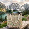 Tote bag with North Cascades National Park design, flannel shirt, map, and water bottle against a mountainous landscape.