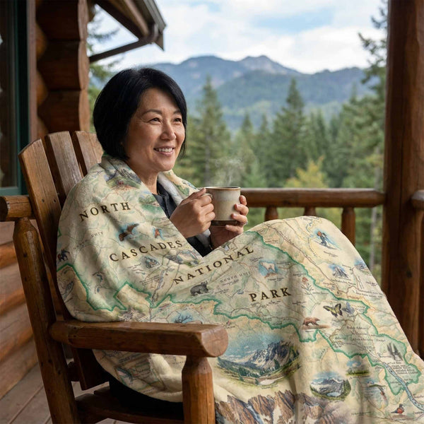 Woman sitting on a wooden chair with a North Cascades National Park blanket, holding a mug, with a scenic background.