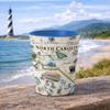 Shot glass with North Carolina map design on a wooden surface with a lighthouse and ocean in the background