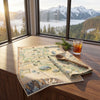 A vintage-style illustrated map laid out on a wooden windowsill with a glass of iced tea, a wooden spoon, and a pine sprig, overlooking a mountain and forest landscape.