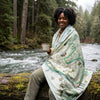 Person sitting on a log by a river, holding a mug and wrapped in a map-themed blanket.