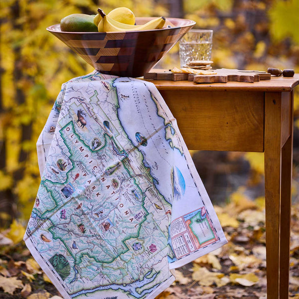 Map blanket draped over a wooden table with a bowl of fruit and glass in an autumn setting