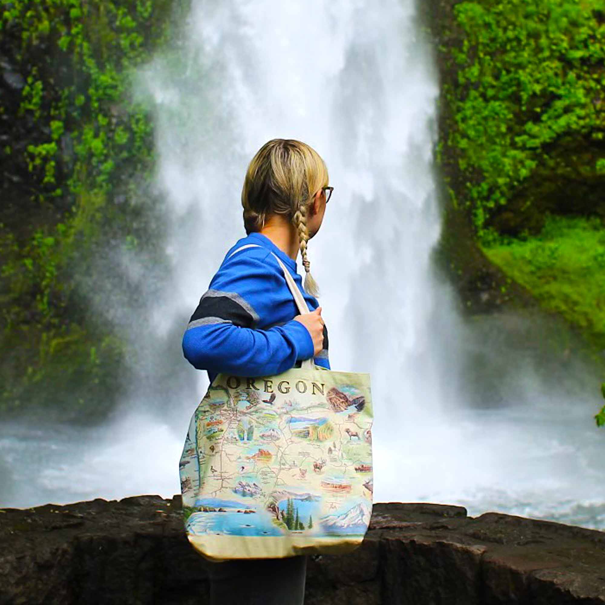 A girl standing next to a waterfall in Oregon carrying a canvas tote bag by Xplorer Maps. 