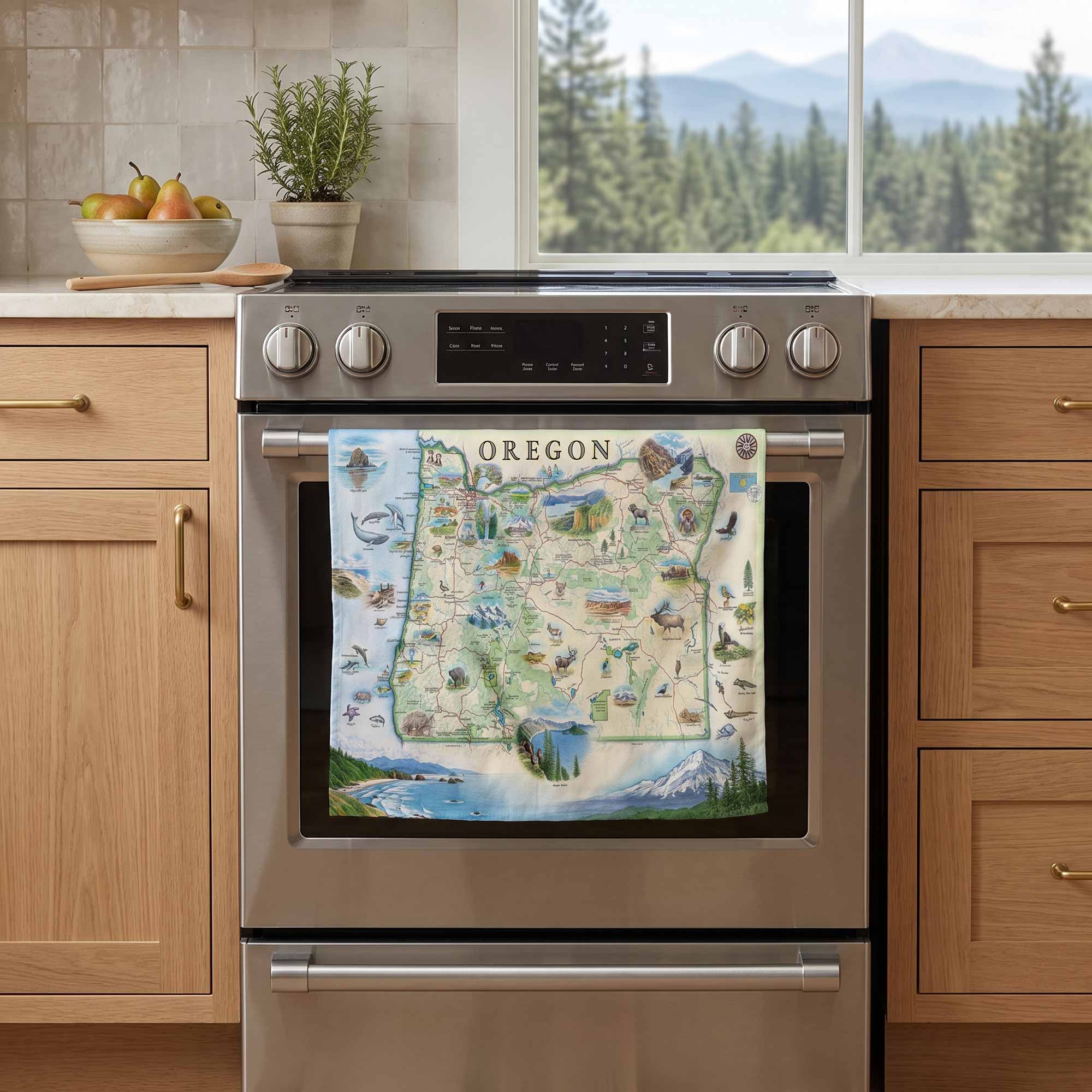 A kitchen scene with a stainless steel oven and a colorful Oregon map print hanging on the oven door. The background shows light wood cabinets, a countertop with fruit and a potted plant, and a window view of trees and mountains.