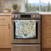 A kitchen scene with a stainless steel oven and a colorful Oregon map print hanging on the oven door. The background shows light wood cabinets, a countertop with fruit and a potted plant, and a window view of trees and mountains.
