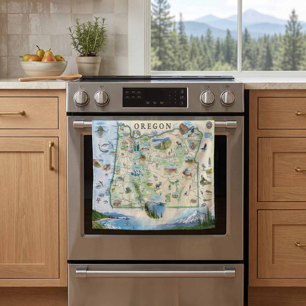 A kitchen scene with a stainless steel oven and a colorful Oregon map print hanging on the oven door. The background shows light wood cabinets, a countertop with fruit and a potted plant, and a window view of trees and mountains.