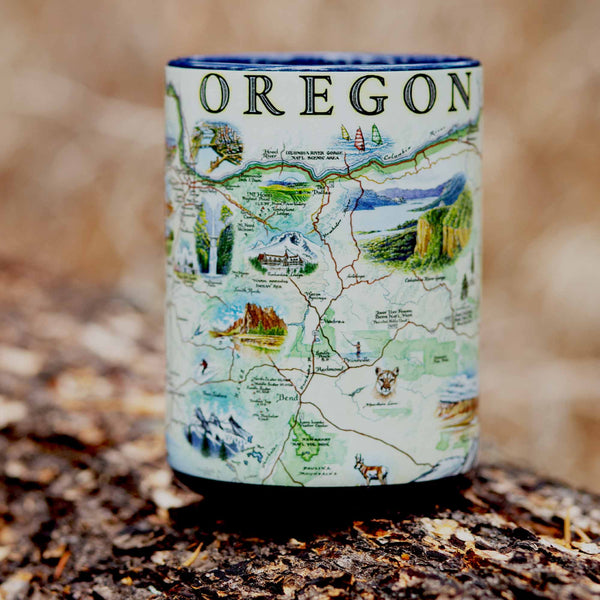Blue 16 oz Oregon State Map Ceramic Mug.  Sitting on a log in the forest. The map features illustrations such as Hells Canyon Snake River, the Columbia River Gorge, Multnomah Falls, and Crater Lake. Flora and fauna include the blue whale, big horn sheep, Dungeness crab, Oregon grape, and Douglas Fir. 