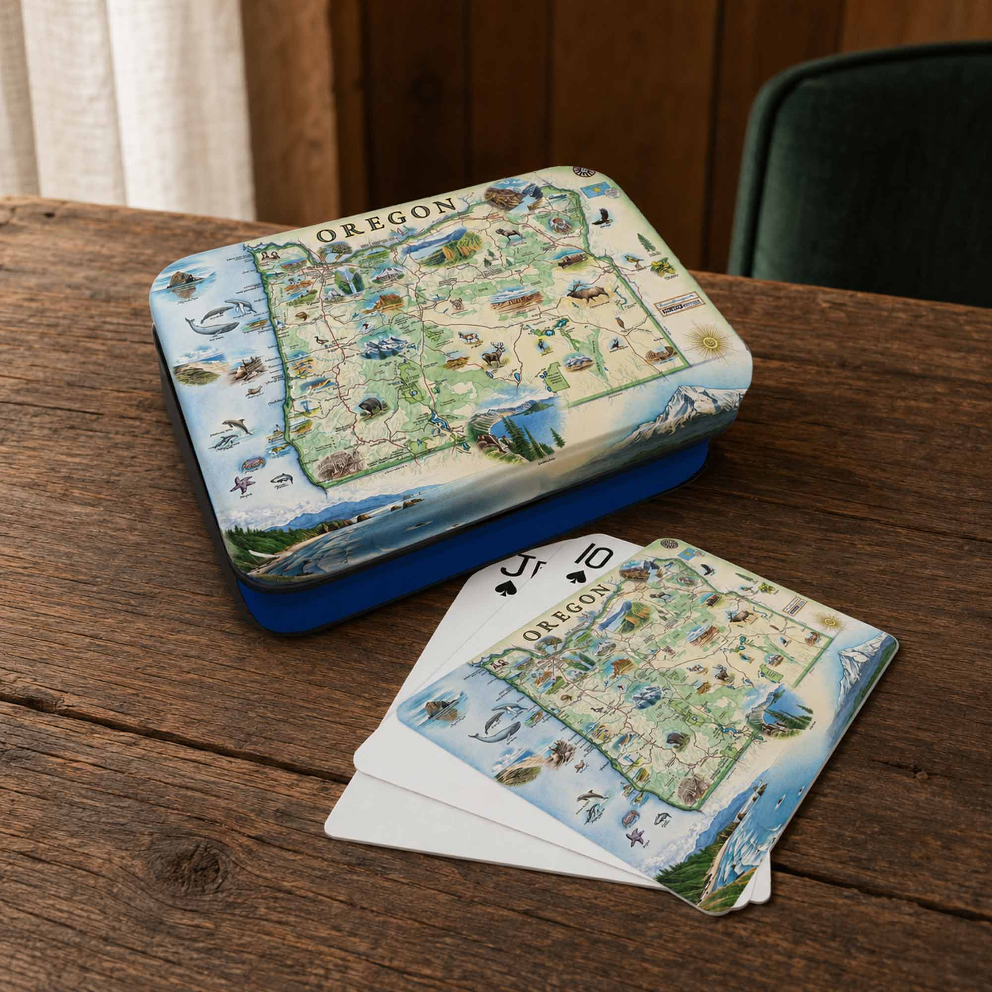 Oregon-themed playing cards and matching tin box displayed on a wooden table. The box lid and card backs feature a vintage-style illustrated map of Oregon with scenic details, and the cards are fanned out in front.