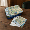 Oregon-themed playing cards and matching tin box displayed on a wooden table. The box lid and card backs feature a vintage-style illustrated map of Oregon with scenic details, and the cards are fanned out in front.