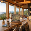 A large Oregon state jigsaw puzzle box and matching puzzle book sit on a wooden dining table in a warm, wood-paneled cabin with large windows overlooking a mountain and lake landscape.
