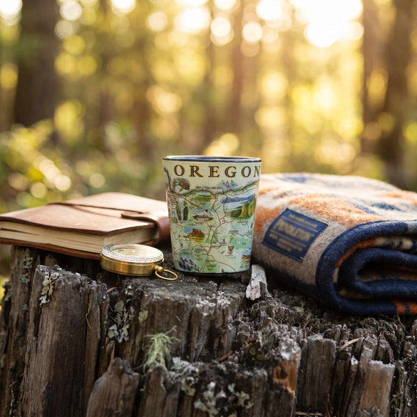 A ceramic mug with a vintage-style Oregon map illustration sits on a rustic tree stump outdoors, surrounded by a notebook, a small compass, and a folded patterned blanket in a sunlit forest setting.